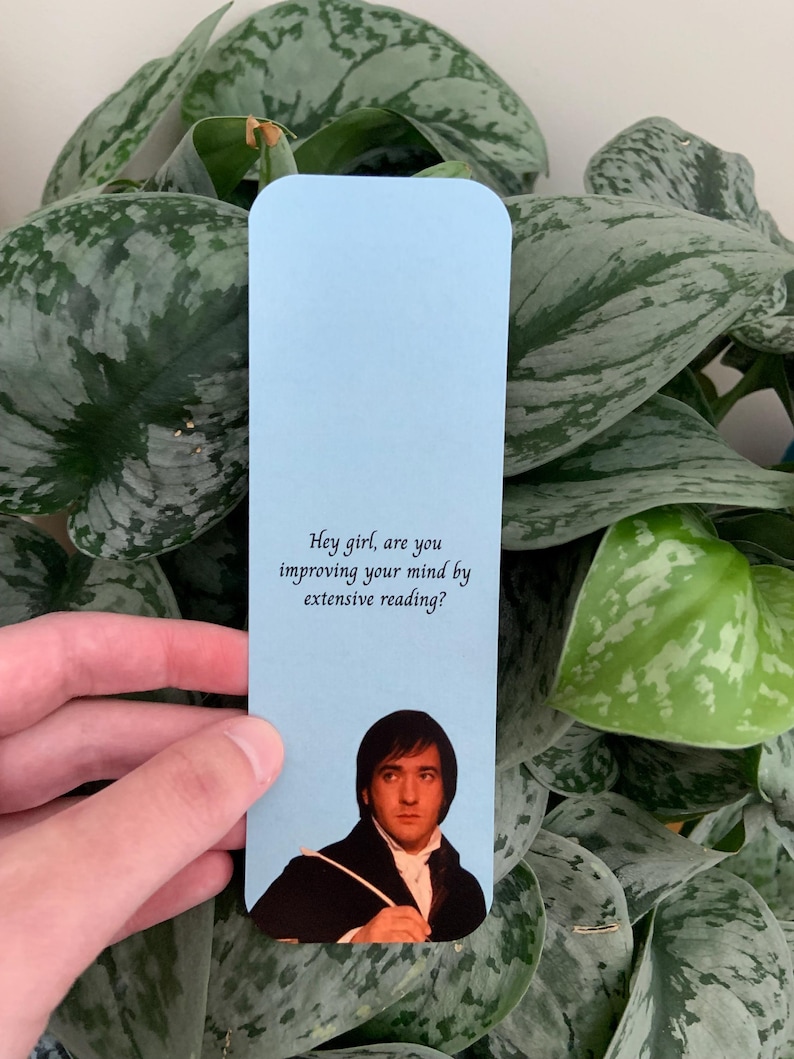 Op de afbeelding: Een lichtblauwe rechthoekige boekenlegger met de gedrukte tekst "Hey girl, are you improving your mind by extensive reading?" boven een fotografische afbeelding van een persoon met donker haar in een zwarte jas en een wit overhemd.