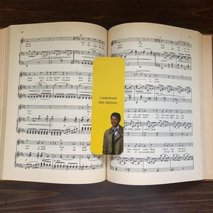 May include: Open book with musical notes and a yellow bookmark. The bookmark has the text "I understand that reference" and a picture of a man. The book is on a dark brown wooden surface.