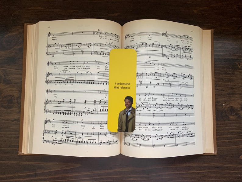 May include: Open book with musical notes and a yellow bookmark. The bookmark has the text "I understand that reference" and a picture of a man. The book is on a dark brown wooden surface.
