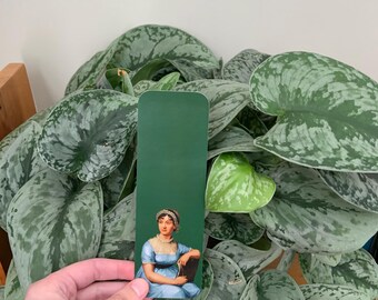 May include: A rectangular bookmark with a solid dark green top section and a bottom section featuring a portrait of a person wearing a light blue dress with a white ruffled collar and a white headpiece, seated while holding a dark book.