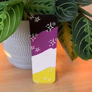 Puede incluir: Marcapáginas con el diseño de la bandera del orgullo no binario. Presenta una parte superior negra, una sección media morada, una franja blanca y una base amarilla. Gráficos de estrellas y estrellas blancas están dispersos por el diseño. El marcapáginas es rectangular.