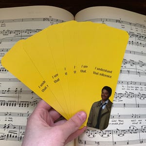 May include: A hand holds a fan of yellow cards with the text "I understand that reference." A small image of a man in a trench coat is on one of the cards. The cards are in front of an open book of sheet music.