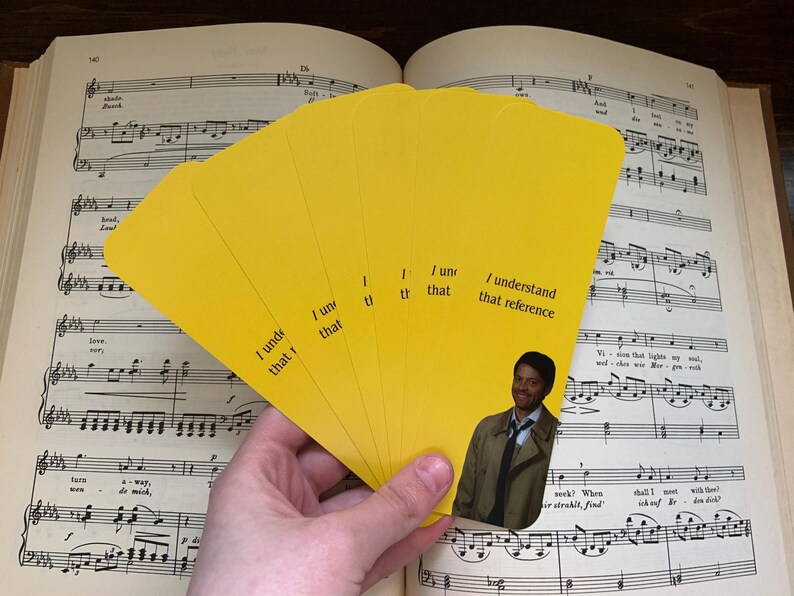 May include: A hand holds a fan of yellow cards with the text "I understand that reference." A small image of a man in a trench coat is on one of the cards. The cards are in front of an open book of sheet music.