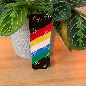 May include: A rectangular bookmark with a black background and colorful diagonal stripes of red, yellow, white, blue, and green. White starburst and star designs are scattered across the black sections.