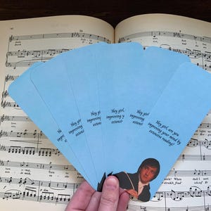 Op de afbeelding: Een waaier van lichtblauwe bladwijzers met een afbeelding van een man in een donkere jas en de tekst, Hey girl, are you improving your mind by extensive reading? De bladwijzers zijn in een halve cirkel over een muziekboek heen geplaatst.