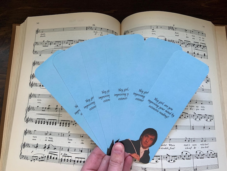 Op de afbeelding: Een waaier van lichtblauwe bladwijzers met een afbeelding van een man in een donkere jas en de tekst, Hey girl, are you improving your mind by extensive reading? De bladwijzers zijn in een halve cirkel over een muziekboek heen geplaatst.