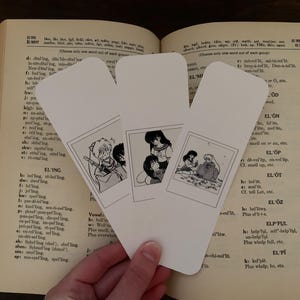 May include: Three white bookmarks with black and white illustrations of anime characters. The bookmarks are on top of an open book.