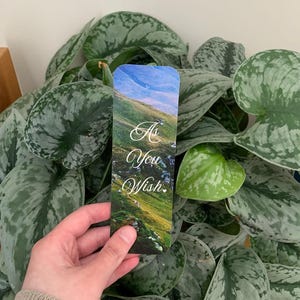 Puede incluir: Un marcapáginas rectangular con una fotografía de paisaje de una colina verde y cubierta de hierba bajo un cielo azul. El texto "As You Wish." está impreso en una elegante letra cursiva blanca en el centro del marcapáginas sobre el paisaje.