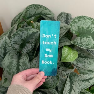 Puede incluir: Un marcapáginas rectangular con un fondo degradado en azul y turquesa que presenta el texto en blanco Don't touch my Dam Book. El marcapáginas tiene un acabado liso y esquinas redondeadas, mostrando una fuente clara y negrita centrada al frente.