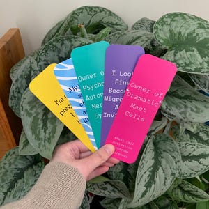 May include: A set of five rectangular bookmarks in yellow, blue with white waves, teal, purple, and hot pink. Each bookmark features white text related to health conditions, including phrases like Owner of Dramatic Mast Cells and #Mast Cell Activation Syndrome Awareness.