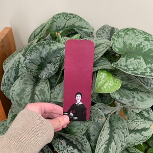 Peut inclure: Un marque-page rectangulaire avec une section supérieure magenta foncé unie et un portrait photographique en noir et blanc d'une personne aux cheveux foncés portant une robe sombre en bas, avec des coins arrondis et une surface lisse.