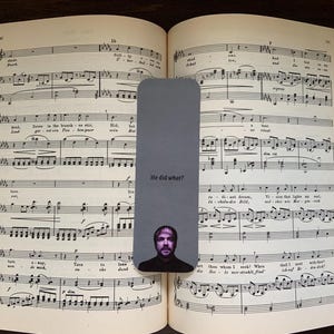 Pode incluir: Um marcador de página cinzento com o texto "He did what?" e um retrato de homem, colocado num livro aberto de partituras. As páginas do livro estão amareladas e o marcador está centrado.