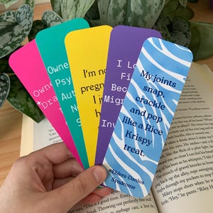 Peut inclure: Un ensemble de cinq marque-pages rectangulaires aux couleurs rose, bleu sarcelle, jaune, violet et bleu avec des motifs ondulés blancs. Les marque-pages comportent du texte imprimé, notamment la phrase 'My joints snap, crackle and pop like a Rice Krispy treat' et '#Ehlers-Danlos Syndrome'.