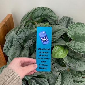 May include: A rectangular blue bookmark featuring a cartoon illustration of a salt container at the top and the text: What kind of house doesn't have salt? Low sodium freaks! The bookmark has a smooth finish and is displayed against a green plant.