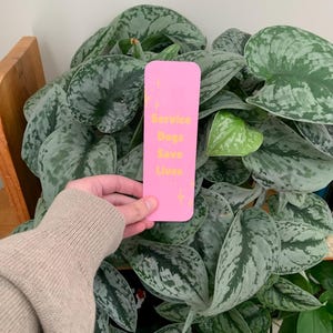 May include: A rectangular pink bookmark with rounded corners featuring the yellow text Service Dogs Save Lives and small yellow star accents. The bookmark is oriented vertically and has a smooth, flat surface finish.
