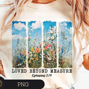 May include: A cream-coloured t-shirt with a colourful floral design set against a blue sky. The design includes the text "LOVED BEYOND MEASURE" and "Ephesians 3:19". The image depicts a meadow with various flowers.
