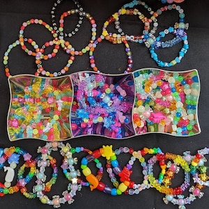 Könnte beinhalten: Eine Sammlung bunter Perlenarmbänder mit verschiedenen Designs, darunter Sterne, Herzen und Buchstaben. Die Armbänder werden auf einem rosa Tablett mit weißem Hintergrund präsentiert.