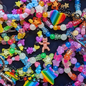 Könnte beinhalten: Eine bunte Auswahl an Perlenketten und Armbändern mit Stern-, Herz- und Blumenanhängern. Eine kleine Plastikpuppe ist in der Mitte des Bildes enthalten.