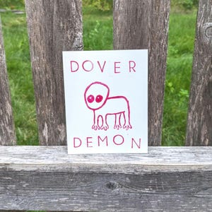 May include: A white card with the words "Dover Demon" printed in pink. A pink cartoon creature with two eyes and four legs is drawn on the card.