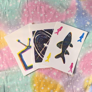 May include: Three art prints on white paper. One print features a colorful TV, another a swirling galaxy, and the third a dark fish with colorful accents. The background is a pastel fabric with star patterns.