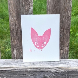 May include: Pink silhouette of a cat's face with white eyes, printed on a white card.