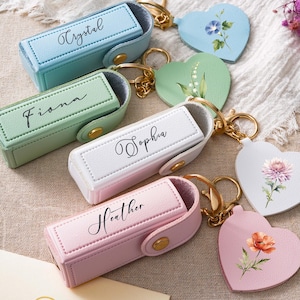 Può includere: Quattro astucci rettangolari per rossetto con chiusure a scatto dorate, ognuno con un nome in corsivo nero: Crystal su azzurro, Fiona su verde salvia, Sophia su bianco e Heather su rosa chiaro. Ogni astuccio include un portachiavi a forma di cuore.