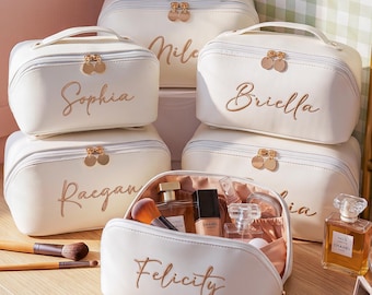 May include: A collection of five cream-colored cosmetic bags with gold-toned zippers and handles. Each bag features a name embroidered in cursive script, including Sophia, Briella, Raegan, and Felicity. The open bag reveals makeup brushes and perfume bottles.
