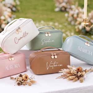 May include: Five rectangular cosmetic bags with top handles and gold-toned double zippers. The bags are white with the name Camille, sage green with Sophia, light blue with MARCIA, pink with AURORA, and tan with Victoria printed on the front.