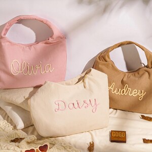 May include: Three corduroy handbags in pink, beige, and brown, each embroidered with a name: Olivia, Daisy, and Audrey. The bags have a rounded shape with a single handle. The bags are on a white surface.