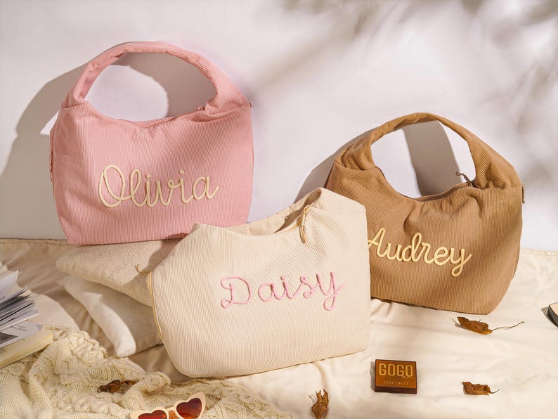 May include: Three corduroy handbags in pink, beige, and brown, each embroidered with a name: Olivia, Daisy, and Audrey. The bags have a rounded shape with a single handle. The bags are on a white surface.