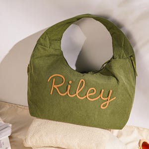 May include: Olive green corduroy shoulder bag with the name "Riley" embroidered in a light orange script. The bag has a curved shape, a zipper, and a single shoulder strap. The bag is resting on a white towel.