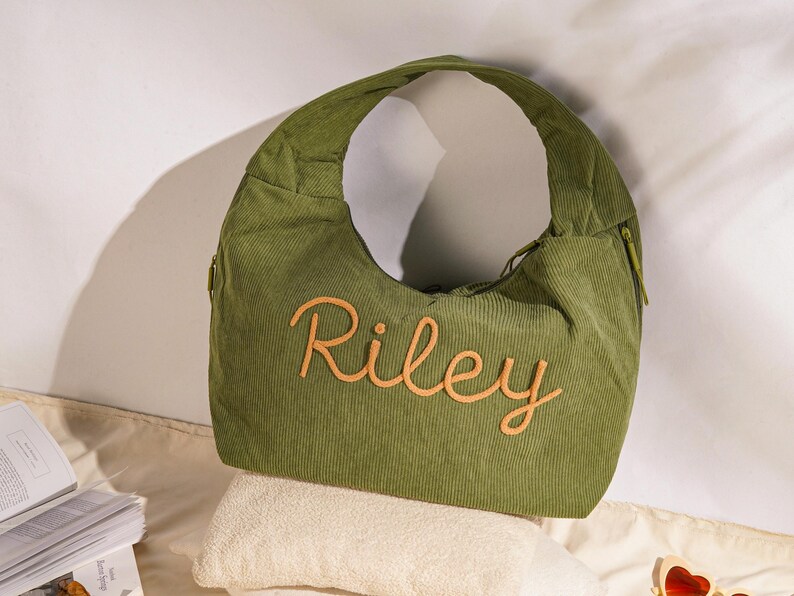 May include: Olive green corduroy shoulder bag with the name "Riley" embroidered in a light orange script. The bag has a curved shape, a zipper, and a single shoulder strap. The bag is resting on a white towel.