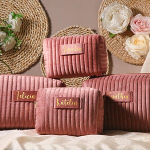 Peut inclure: Cinq trousses de maquillage matelassées roses avec des lettres dorées. Les trousses sont étiquetées "Felicia", "Katelyn", "Amelia", "Smitha" et "Felicia".