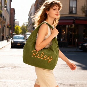 May include: Olive green tote bag with the name "Riley" embroidered in orange script. The bag is carried over the shoulder. The image is taken outdoors on a city street.