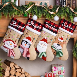 Op de afbeelding: Vier kerstsokken van jute met een rood-zwart geruite boord en wit geborduurde namen: Bernd, Kayla, Chloe en Sophia. Elke sok is voorzien van een pluche figuur: een sneeuwpop, pinguïn, de kerstman en een rendier, elk met een sjaal om.