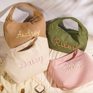 May include: Four corduroy shoulder bags in tan, olive green, cream, and pink, each embroidered with a name: Audrey, Riley, Daisy, and Olivia. The bags have a curved shape and a single handle. Sunglasses and a small cosmetic item are also visible.
