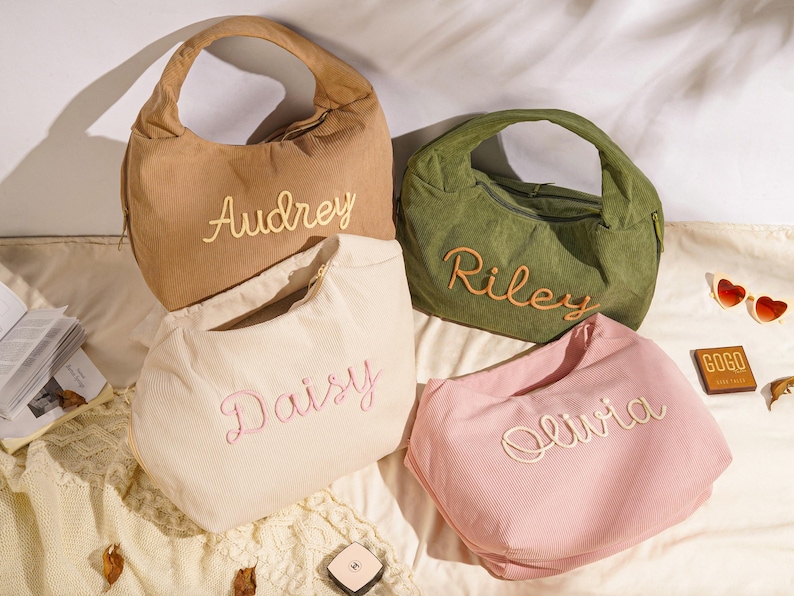 May include: Four corduroy shoulder bags in tan, olive green, cream, and pink, each embroidered with a name: Audrey, Riley, Daisy, and Olivia. The bags have a curved shape and a single handle. Sunglasses and a small cosmetic item are also visible.
