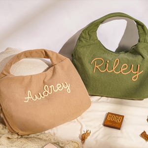 May include: Two corduroy shoulder bags, one tan and one olive green, each with a name embroidered in a contrasting color. The tan bag says "Audrey" and the green bag says "Riley". A small wooden box with the text "GOGO" is also visible.