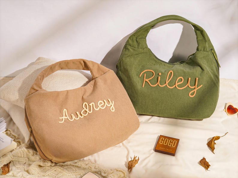 May include: Two corduroy shoulder bags, one tan and one olive green, each with a name embroidered in a contrasting color. The tan bag says "Audrey" and the green bag says "Riley". A small wooden box with the text "GOGO" is also visible.