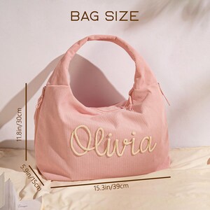 May include: Pink corduroy shoulder bag with the name "Olivia" embroidered on the front. The bag is 15.3 inches wide, 11.8 inches high, and 5.9 inches deep. The bag has a large handle and a zipper closure.