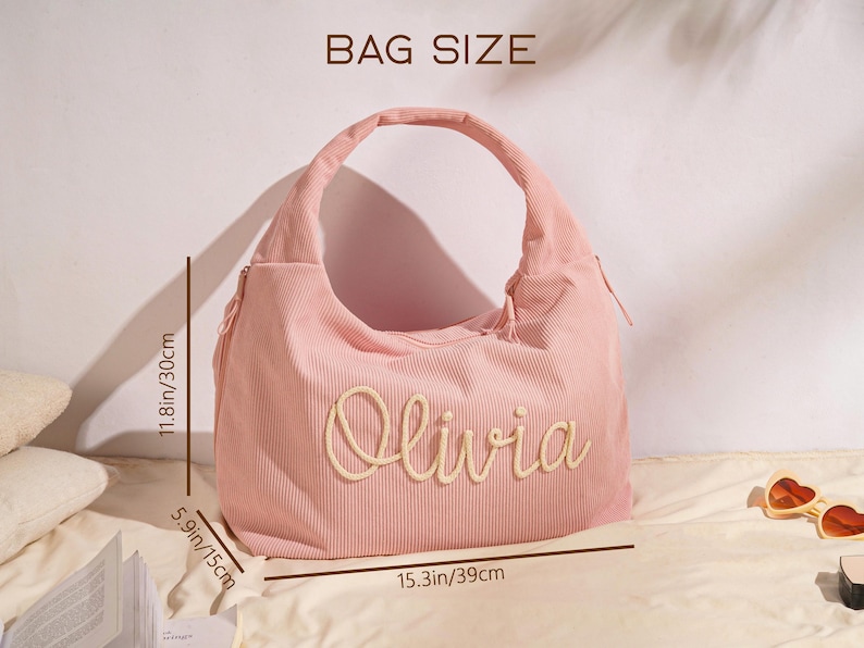 May include: Pink corduroy shoulder bag with the name "Olivia" embroidered on the front. The bag is 15.3 inches wide, 11.8 inches high, and 5.9 inches deep. The bag has a large handle and a zipper closure.