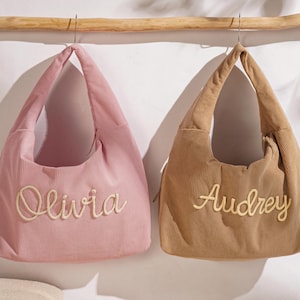 May include: Two corduroy tote bags, one pink and one tan, hang from a wooden rod. The pink bag has "Olivia" embroidered on it, and the tan bag has "Audrey" embroidered on it. Both bags have a simple, rounded shape.