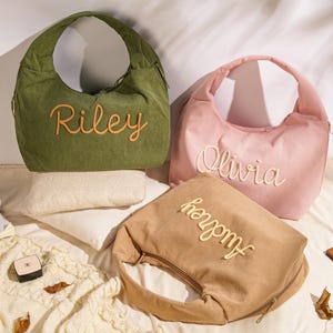 May include: Three corduroy shoulder bags in olive green, pink, and tan, each embroidered with a name in a contrasting color. The bags have a crescent shape and a zipper closure. The names embroidered are Riley, Olivia, and Audrey.