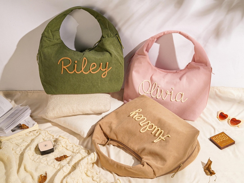 May include: Three corduroy shoulder bags in olive green, pink, and tan, each embroidered with a name in a contrasting color. The bags have a crescent shape and a zipper closure. The names embroidered are Riley, Olivia, and Audrey.