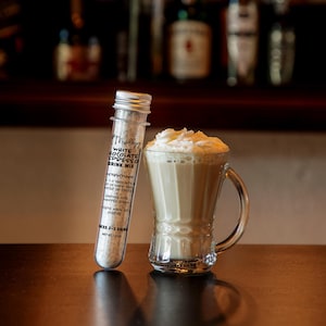 Może przedstawiać: Przezroczysta plastikowa prob&oacute;wka wypełniona mieszanką do napoju espresso z białą czekoladą, ze srebrną zakrętką i etykietą z instrukcją. Obok znajduje się szklany kubek z kremowym napojem w kolorze beżowym, udekorowany białą bitą śmietaną.