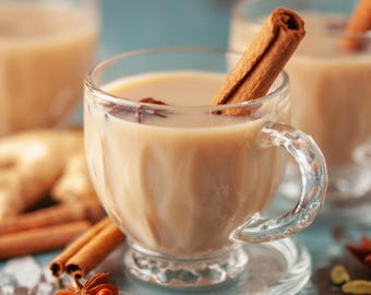May include: A close-up of a glass cup filled with a creamy, light brown beverage, likely chai tea. A cinnamon stick and star anise are visible in the drink. Other spices and ingredients, including cardamom and peppercorns, are scattered around the cup on a light blue surface.
