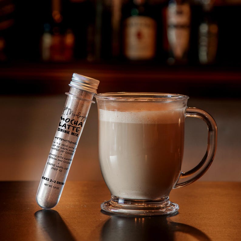 Może przedstawiać: Przezroczysta plastikowa prob&oacute;wka wypełniona mieszanką do przygotowania latte mokka ze srebrną zakrętką i etykietą z napisem Mocha Latte Drink Mix, obok szklanego kubka zawierającego jasnobrązowy, kremowy nap&oacute;j kawowy.