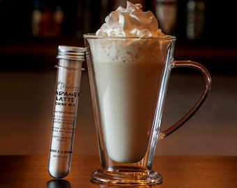 May include: A clear glass test tube containing caramel latte drink mix with a silver screw cap, positioned next to a tall glass mug filled with a creamy beige latte topped with a generous swirl of white whipped cream. The label reads Caramel Latte.
