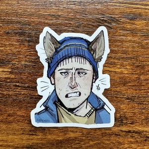 May include: A sticker featuring a cartoon illustration of a person wearing a blue beanie with dog ears. The person has an angry expression, with furrowed brows and a scowl. The sticker is on a wooden surface.