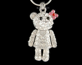 May include: A silver necklace with a crystal-encrusted teddy bear charm. The bear has a pink flower on its head with red and pink crystals.